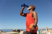 Ajuste o homem americano africano sênior pelo mar que bebe da garrafa de água que prende o basquete. esporte de aposentadoria saudável ao ar livre fitness lifestyle. — Fotografia de Stock