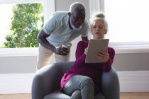 Senioren-Mixed-Race-Paar sitzt auf der Couch und schaut gemeinsam auf das digitale Tablet im Wohnzimmer. Mann trinkt Kaffee. Während der Quarantäne zu Hause bleiben und sich selbst isolieren. — Stockfoto