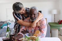 Pareja afroamericana mayor sentada a la mesa cenando y abrazándose en casa. senior retiro estilo de vida amigos socializar. - foto de stock