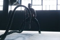 Uomo afroamericano che indossa vestiti sportivi combattendo corde in un edificio urbano vuoto. fitness urbano stile di vita sano. — Foto stock