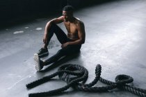 Afroamerikanischer Mann in Sportkleidung sitzt nach einem Kampf mit Seilen in einem leeren städtischen Gebäude und ruht. urbane Fitness gesunder Lebensstil. — Stockfoto