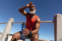 Подходит старший африканский американец, занимающийся спортом, сидя на солнце, держа бутылку с водой. здоровый отдых фитнес на открытом воздухе. — стоковое фото