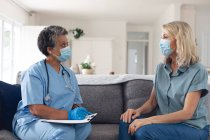 Doctora afroamericana mayor hablando con paciente femenina ambas usando mascarillas en casa. protección de la higiene sanitaria durante la pandemia del coronavirus covid 19. - foto de stock