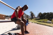 Подходит старший африканский американец, занимающийся спортом, сидя на солнце, держа бутылку с водой. здоровый отдых фитнес на открытом воздухе. — стоковое фото