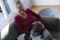 Старшая пара смешанных рас обнимается, глядя на смартфон в гостиной. оставаться дома в изоляции во время карантинной изоляции. — стоковое фото