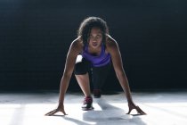Mujer afroamericana vestida con ropa deportiva arrodillada empezando a correr en un edificio urbano vacío. aptitud urbana estilo de vida saludable. - foto de stock