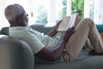Homme afro-américain aîné allongé sur un canapé lisant un livre. rester à la maison en isolement personnel pendant le confinement en quarantaine. — Photo de stock