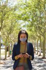 Mujer afroamericana con máscara facial usando smartphone en la calle. estilo de vida durante la pandemia de coronavirus covid 19. - foto de stock