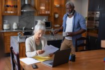 Senior mixed race couple using laptop paying bills together in dining room. staying at home in self isolation during quarantine lockdown. — Stock Photo