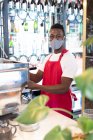 Ritratto di barista afroamericano di sesso maschile che indossa una maschera facciale con macchina da caffè guardando la macchina fotografica. salute e igiene negli affari durante la pandemia di coronavirus covid 19. — Foto stock