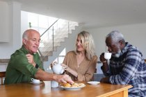 Personas mayores caucásicas y afroamericanas sentadas a la mesa tomando té en casa. senior retiro estilo de vida amigos socializar. - foto de stock