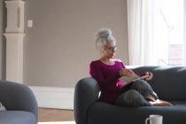 Старшая белая женщина, сидящая на диване в гостиной и читающая книгу. оставаться дома в изоляции во время карантинной изоляции. — стоковое фото