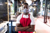 Портрет африканського чоловічого бариста, одягненого в маску обличчя, дивиться на камеру. здоров'я та гігієна в бізнесі під час коронавірусної ковини 19 пандемії. — стокове фото