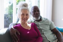 Senior mixed race couple embracing looking at the camera and smiling in living room. staying at home in self isolation during quarantine lockdown. — Stock Photo