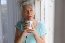 Mujer caucásica mayor de pie junto a la ventana bebiendo una taza de café en casa. permanecer en casa en aislamiento durante el bloqueo de cuarentena. - foto de stock