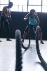 Mulher afro-americana vestindo roupas esportivas batalhando cordas em prédio urbano vazio. O homem está a animá-la. fitness urbano estilo de vida saudável. — Fotografia de Stock