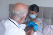 Médico varón caucásico mayor vacunando a una paciente femenina con máscaras faciales en casa. protección de la higiene sanitaria durante la pandemia del coronavirus covid 19. - foto de stock