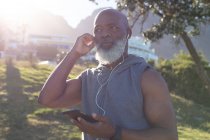 Fit senior homme afro-américain tenant smartphone mettre des écouteurs. saine retraite technologie communication extérieur forme physique mode de vie. — Photo de stock
