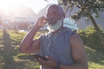 Ajuste homem americano africano sênior segurando smartphone colocando fones de ouvido. tecnologia de aposentadoria saudável comunicação ao ar livre fitness lifestyle. — Fotografia de Stock