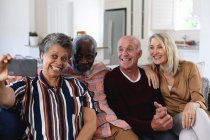 Casais caucasianos e afro-americanos sênior sentados no sofá tirando uma selfie em casa. sênior aposentadoria estilo de vida amigos socialização. — Fotografia de Stock
