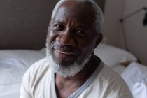 Портрет старшего африканского американца, сидящего на кровати, смотрящего в камеру и улыбающегося. оставаться дома в изоляции во время карантинной изоляции. — стоковое фото