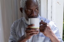 Senior african american man standing by window drinking coffee at home. staying at home in self isolation during quarantine lockdown. — Stock Photo