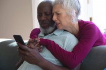 Senioren-Mixed-Race-Paar umarmen sich beim gemeinsamen Blick auf das Smartphone im Wohnzimmer. Während der Quarantäne zu Hause bleiben und sich selbst isolieren. — Stockfoto