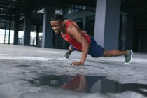 Uomo afroamericano che indossa vestiti sportivi facendo flessioni con una mano in un edificio urbano vuoto. fitness urbano stile di vita sano. — Foto stock