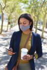 Африканська американка, одягнена в маску обличчя з чашкою кави, використовує смартфон на вулиці. Життя під час коронавірусної ковини 19. — стокове фото