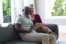 Senior mixed race couple sitting on couch looking at digital tablet together in living room. staying at home in self isolation during quarantine lockdown. — Stock Photo