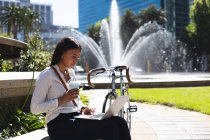 Африканська американка з чашкою кави за допомогою ноутбука сидячи в корпоративному парку. Концепція життя під час коронавірусної ковини 19. — стокове фото