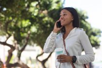 Mujer afroamericana con taza de café hablando en smartphone en la calle. estilo de vida concepto de vida durante el coronavirus covid 19 pandemia. - foto de stock