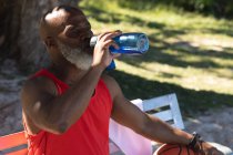 Ajuste o homem americano africano sênior que está sentado no parque que bebe da garrafa de água. esporte de aposentadoria saudável ao ar livre fitness lifestyle. — Fotografia de Stock