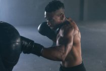 Hombre afroamericano con guantes de boxeo golpeando saco de boxeo en un edificio urbano vacío. aptitud urbana estilo de vida saludable. - foto de stock