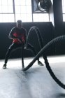 Hombre afroamericano con ropa deportiva luchando contra cuerdas en un edificio urbano vacío. aptitud urbana estilo de vida saludable. - foto de stock