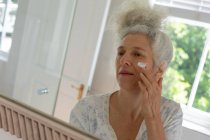 Eine ältere kaukasische Frau steht im Badezimmer und cremt ihr Gesicht ein. Während der Quarantäne zu Hause bleiben und sich selbst isolieren. — Stockfoto
