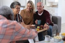 Senior caucasian and african american couples sitting by table eating cheese and fruits at home. senior retirement lifestyle friends socializing. — Stock Photo