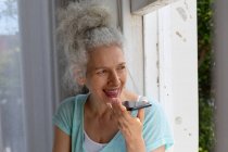 Старша біла жінка стоїть біля вікна, розмовляючи по смартфонах вдома. перебування вдома в самоізоляції під час карантину . — стокове фото
