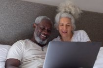 Старшая смешанная раса пара в спальне лежит на кровати с помощью ноутбука. оставаться дома в изоляции во время карантинной изоляции. — стоковое фото