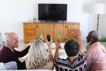 Casais seniores caucasianos e afro-americanos sentados no sofá assistindo ao jogo bebendo cerveja em casa. sênior aposentadoria estilo de vida amigos socialização. — Fotografia de Stock