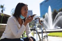 Donna afroamericana con maschera abbassata che parla sullo smartphone mentre è seduta nel parco aziendale. stile di vita concetto di vita durante coronavirus covid 19 pandemia. — Foto stock