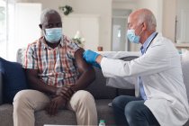 Médecin caucasien principal vaccinant le patient masculin portant tous deux des masques faciaux à la maison. protection de l'hygiène des soins de santé pendant une pandémie de coronavirus covid 19. — Photo de stock