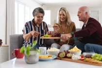 Ältere kaukasische und afrikanisch-amerikanische Paare sitzen zu Hause am Tisch und essen Käse und Obst. Senioren-Lebensstil Freunde Geselligkeit. — Stockfoto