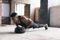 Homem afro-americano exercitando-se fazendo flexões em uma bola de remédios em um prédio urbano vazio. fitness urbano estilo de vida saudável — Fotografia de Stock