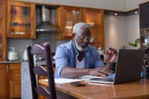 Hombre afroamericano mayor usando laptop pagando facturas en el comedor. permanecer en casa en aislamiento durante el bloqueo de cuarentena. - foto de stock