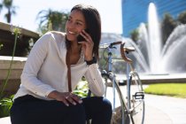 African american woman talking on smartphone while sitting at corporate park. lifestyle living concept during coronavirus covid 19 pandemic. — Stock Photo