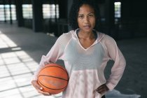Retrato de una mujer afroamericana de pie en un edificio urbano vacío y sosteniendo una pelota de baloncesto. aptitud urbana estilo de vida saludable. - foto de stock