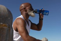 Ajuste hombre afroamericano mayor sentado bebiendo de la botella de agua contra el cielo azul. saludable retiro tecnología comunicación al aire libre fitness estilo de vida. - foto de stock