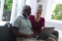 Senioren-Mixed-Race-Paar sitzt auf der Couch und schaut gemeinsam auf Laptop im Wohnzimmer. Während der Quarantäne zu Hause bleiben und sich selbst isolieren. — Stockfoto