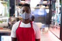 Ritratto di un barista afroamericano di sesso maschile che indossa una maschera che guarda la macchina fotografica. salute e igiene negli affari durante la pandemia di coronavirus covid 19. — Foto stock